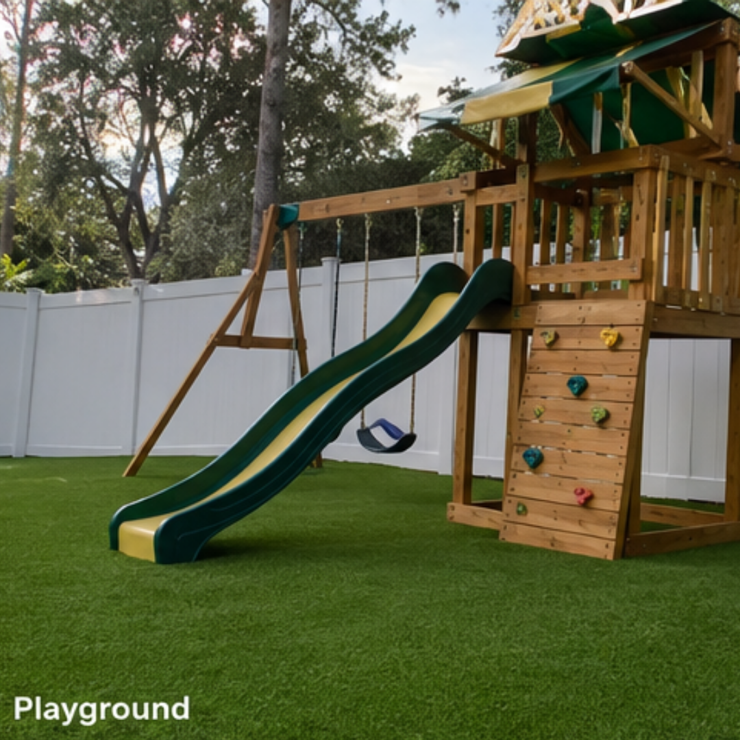Wooden outdoor playground with a green-yellow double slide, a swing set, and a climbing wall in a fenced yard on artificial turf. Includes a shaded platform at top right.