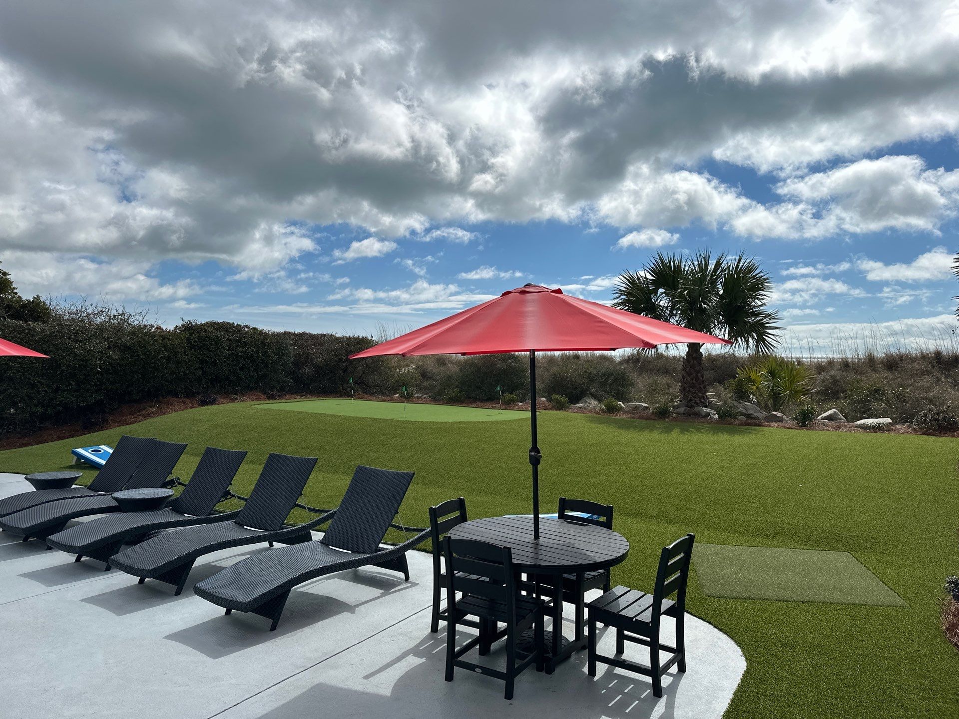 Outdoor poolside seating area with black lounge chairs, a red umbrella, and a round black table on a concrete patio.
