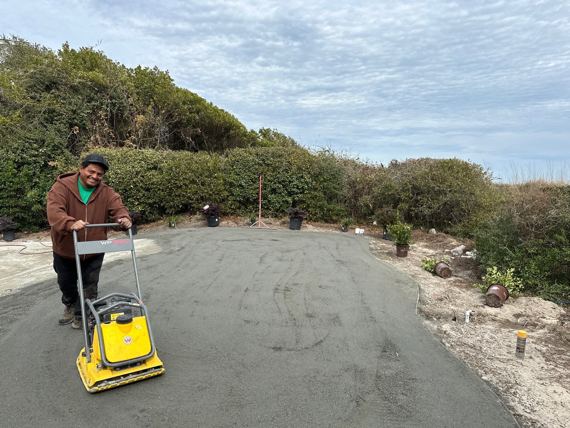 Man in a brown jacket and green shirt operating a yellow plate compactor on a curved paved road with dense shrubs in the background and a cloudy sky.
