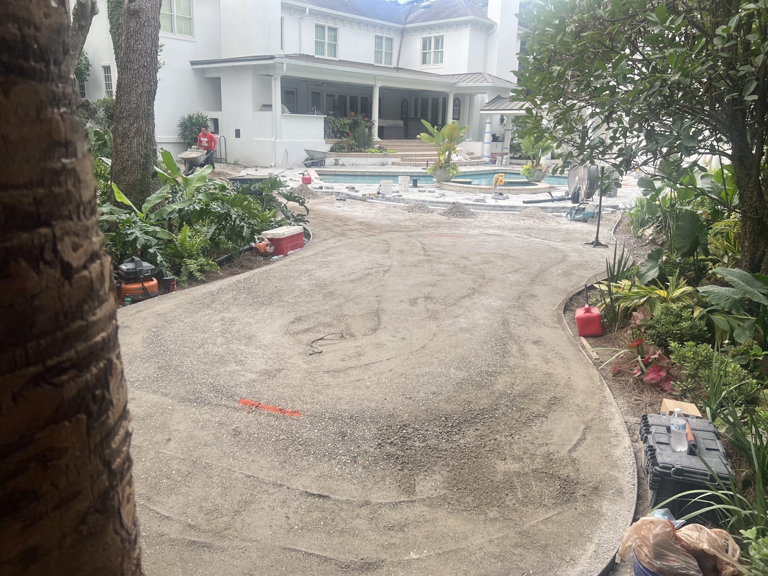 Backyard pool area under renovation with construction materials, tools, and a person in a red shirt near a wheelbarrow in front of a white house.