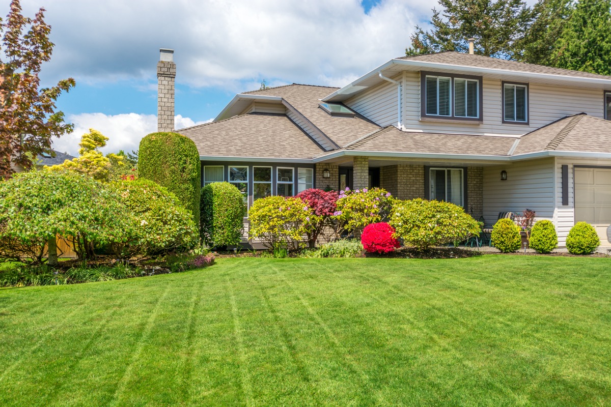 Two-story suburban house with beige siding and a wide front porch, surrounded by a neatly manicured lawn and colorful shrubbery on a sunny day.