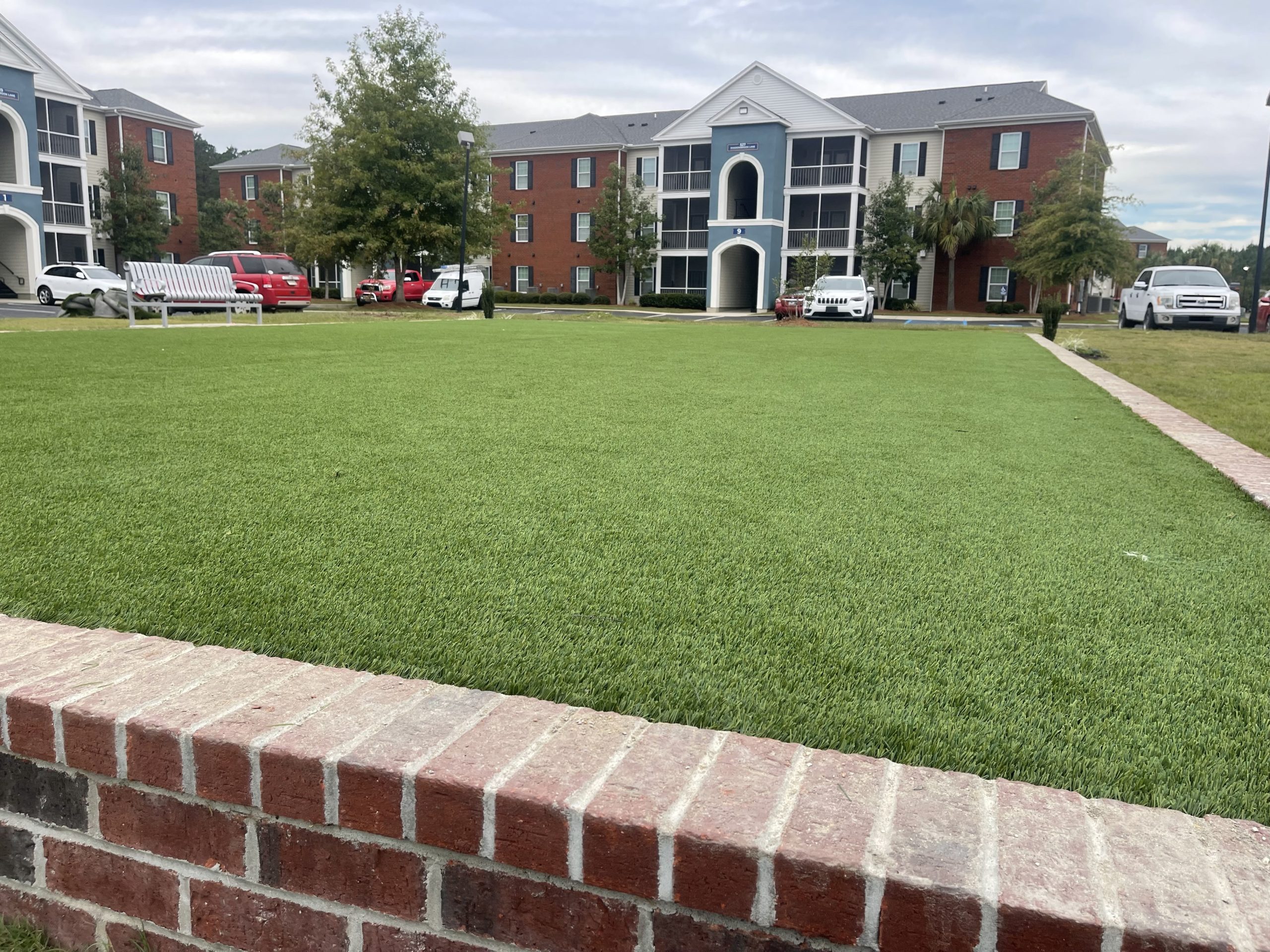 Brick railing in the foreground with a green lawn and multi-story brick apartment buildings in the background, cars parked along the street in a residential complex.