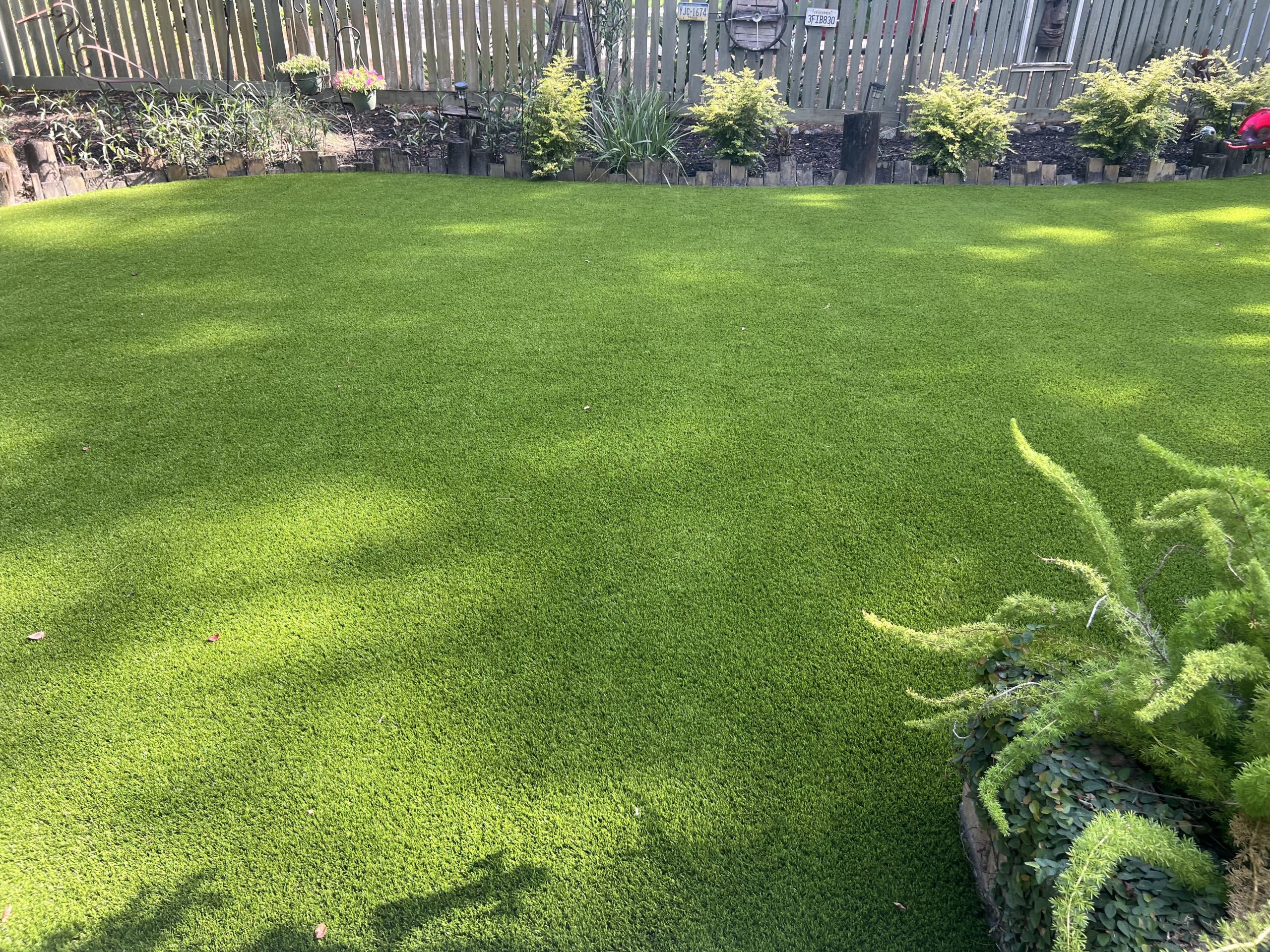 Artificial Grass Header Backyard with a vibrant green artificial lawn and wooden border along a flower bed, fence in the background, and potted plants hanging from the fence.