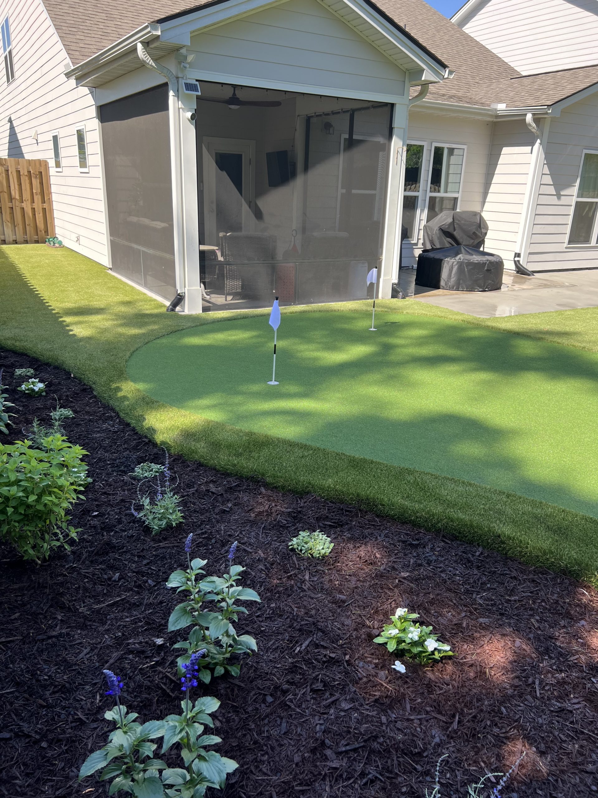 Backyard with screened porch, artificial putting green with two hole flags, and mulched flower beds.