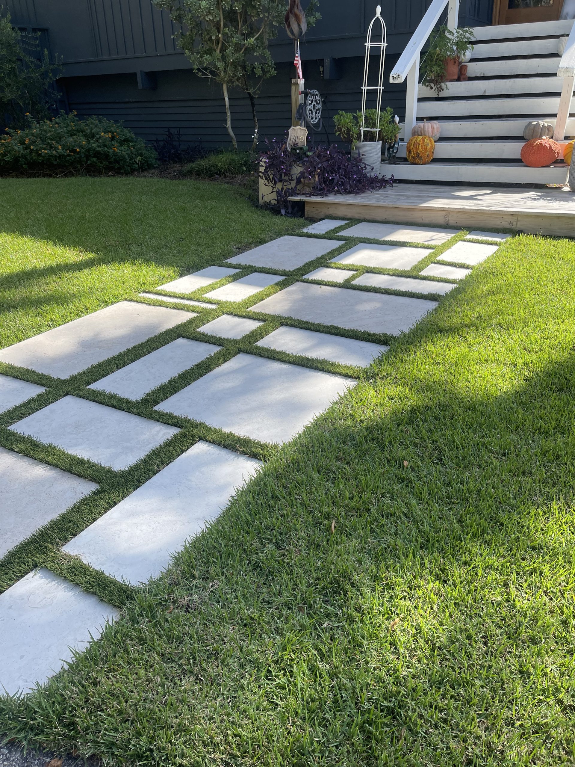Concrete stepping stones set in a green lawn form a path toward a wooden porch with pumpkins on the stairs and potted plants nearby.