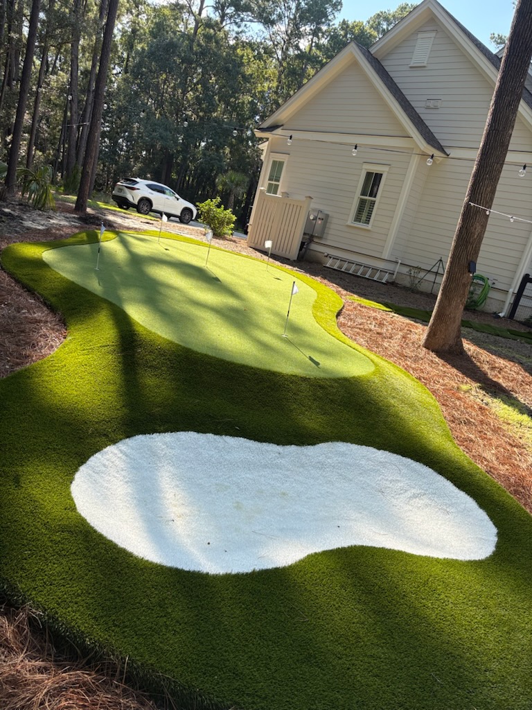 Backyard putting green with a white sand bunker in the foreground, a house and string lights in the background, and a parked white SUV among trees.