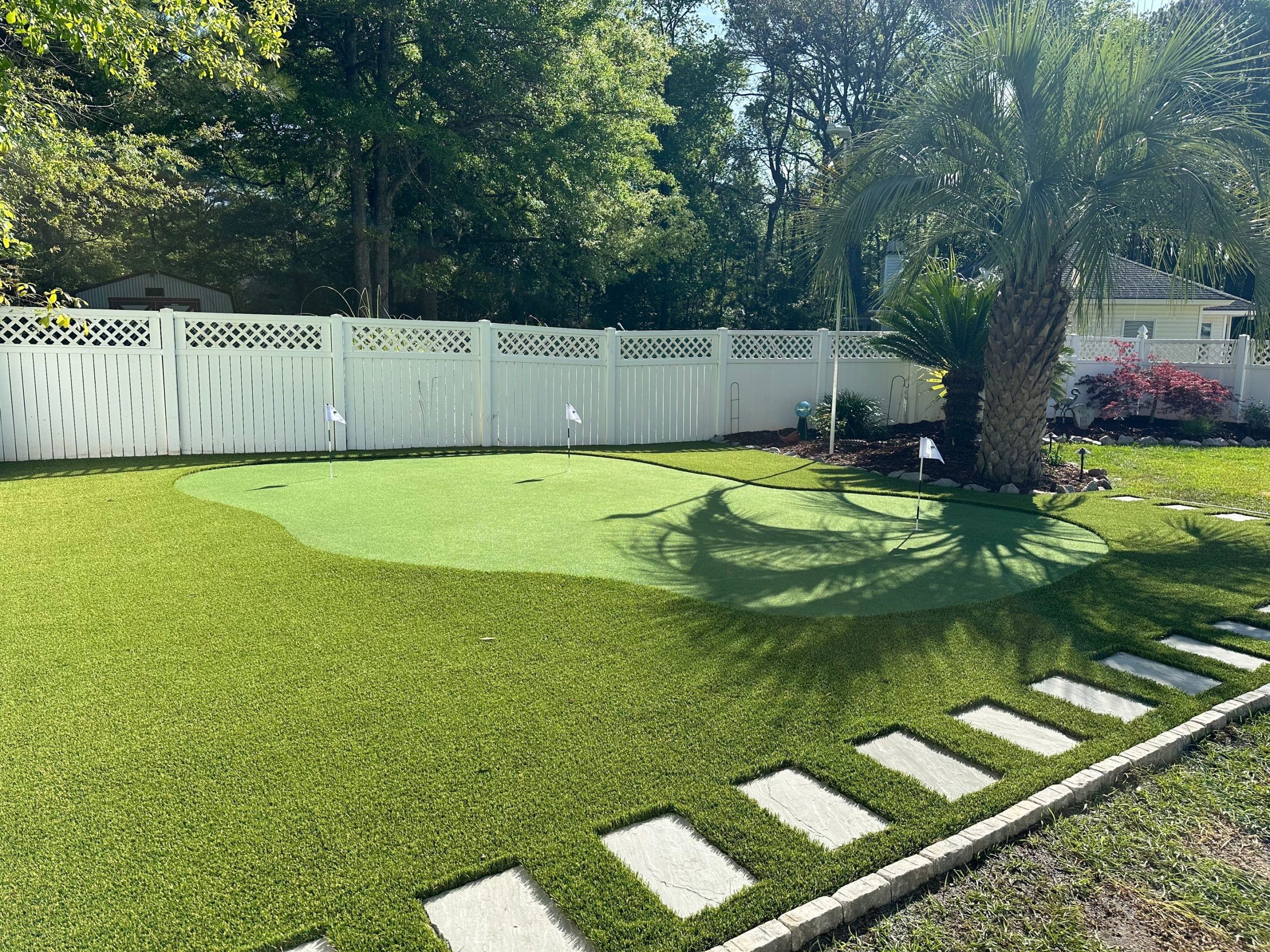 Backyard putting green with white lattice fence, palm tree, and stepping-stone path along the edge of a lawn garden.