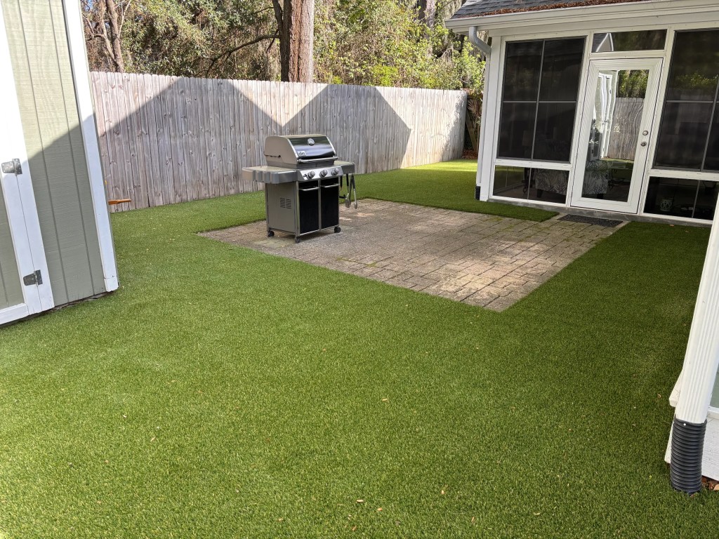 Gas grill on a stone patio in a fenced backyard with artificial grass and a glass-door patio area behind it.