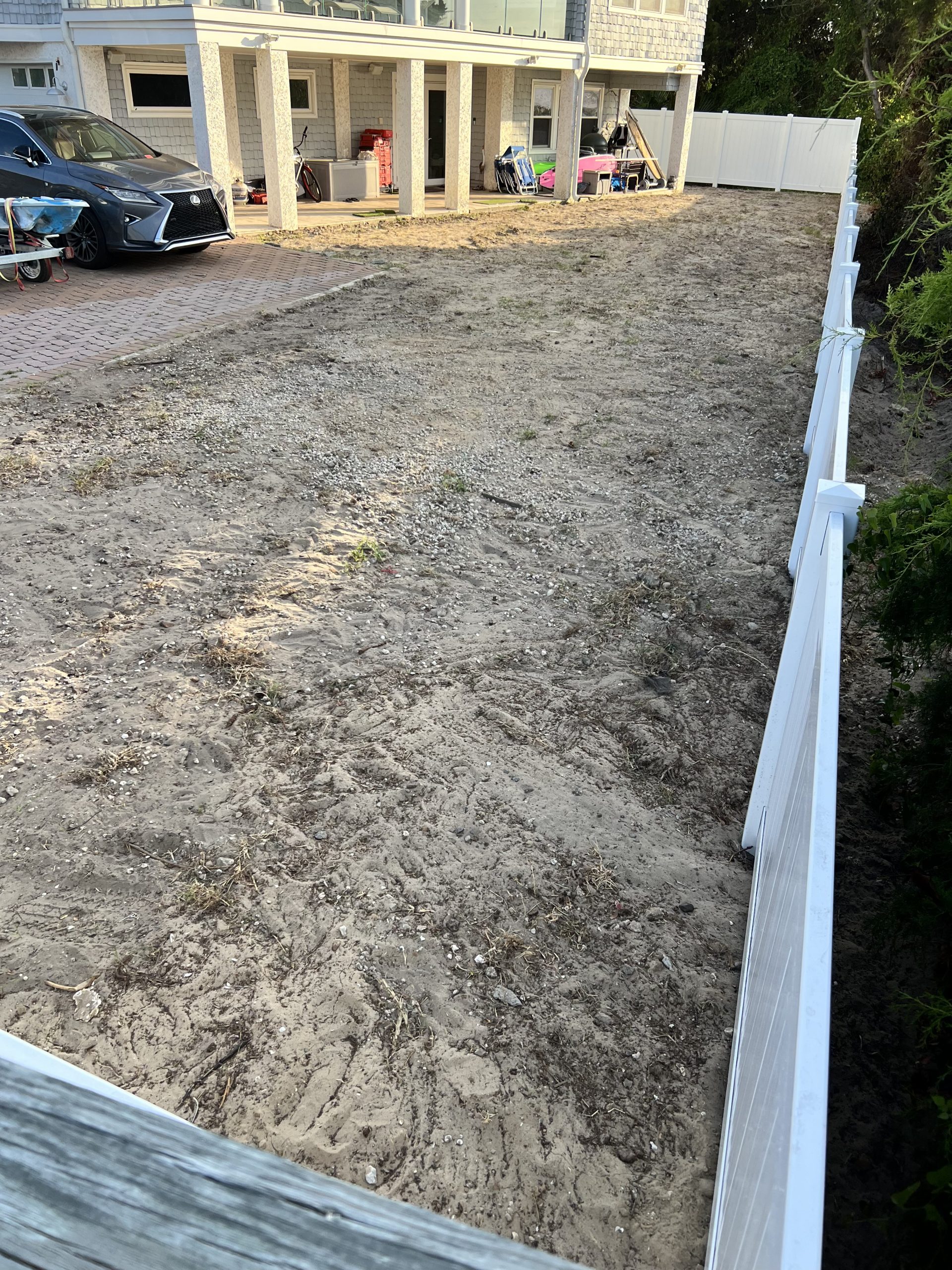 Dirt backyard with a white vinyl fence on the right, a parked gray car and trailer on the left, and a house with a covered patio in the background.