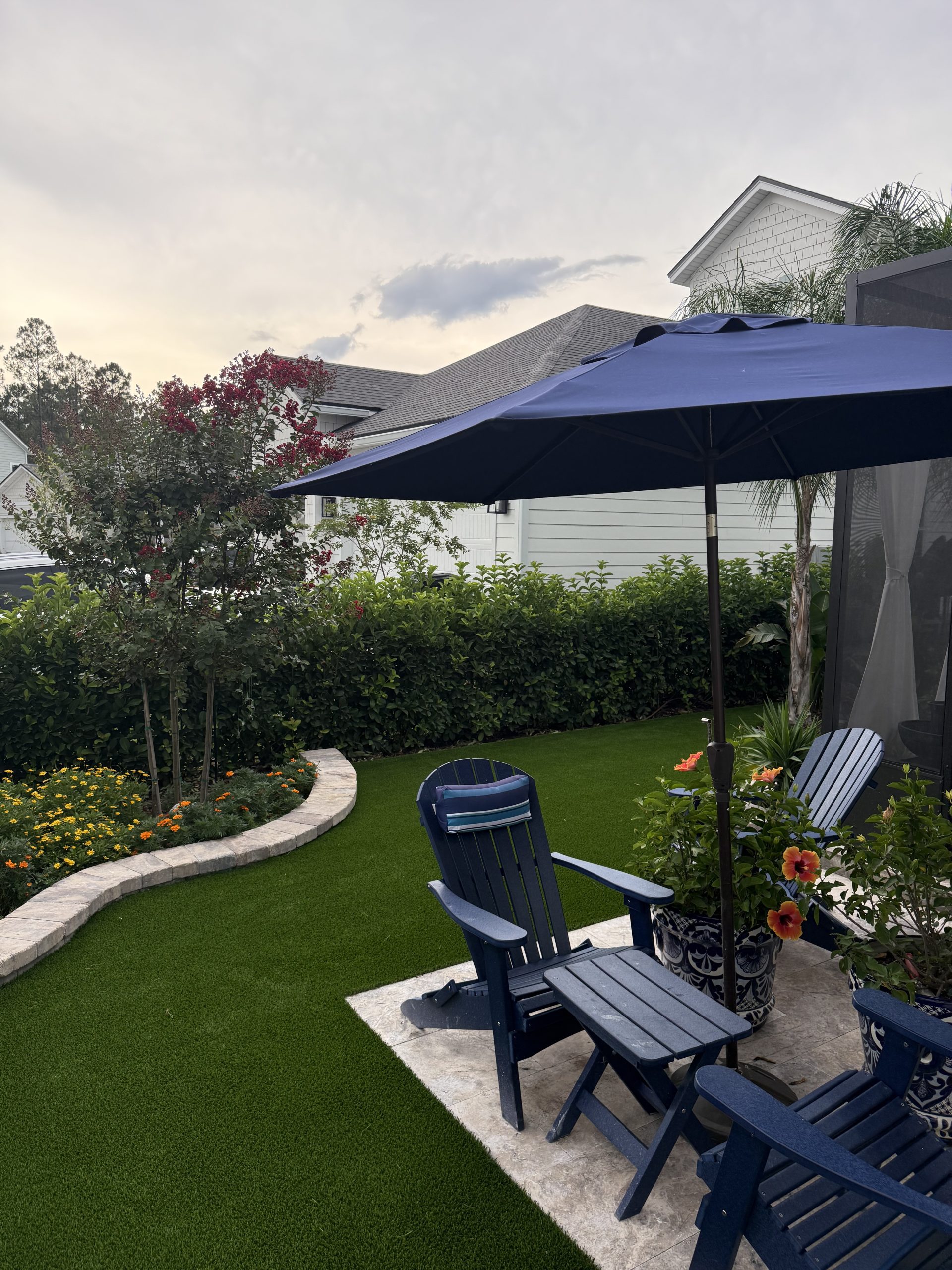 Backyard patio with blue Adirondack chairs under a large navy umbrella, artificial turf, and a flowering bed near a white house.